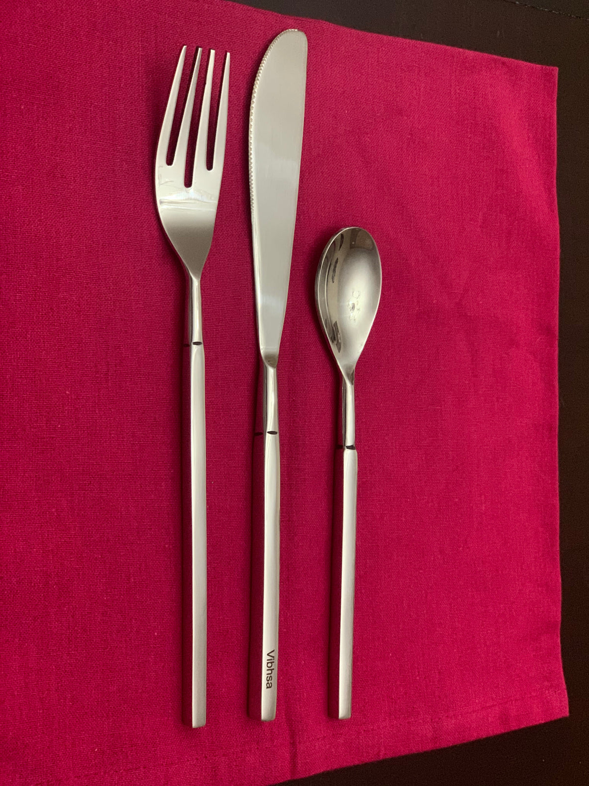 Stainless steel flatware set including a dinner fork, serrated dinner knife, and dessert spoon with straight handles, arranged on a red cloth.
