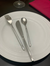Silver stainless steel dinner fork, dinner knife, and dessert spoon on a white round plate with concentric ridge patterns beside a red napkin and a clear glass stemware on a black table.