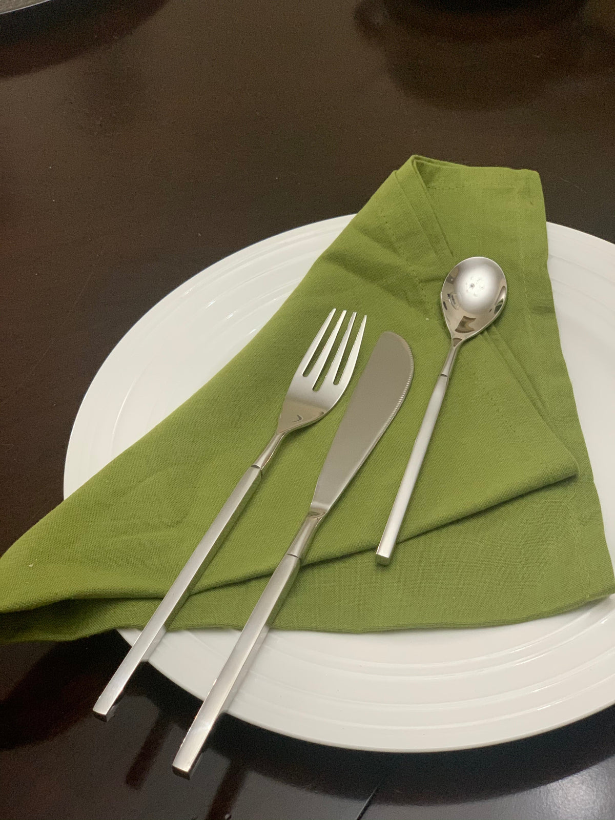 Silver dinner fork, dinner knife, and dessert spoon with squared handles arranged atop a folded olive-green napkin on a white ceramic plate.
