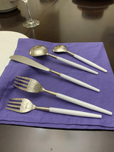 White and silver flatware including two forks, a butter knife, and two spoons, arranged on a purple cloth on a dark wood table.
