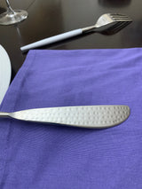 Silver stainless steel knife with textured handle resting on a purple cloth napkin, accompanied by a fork with a white handle on a dark surface.
