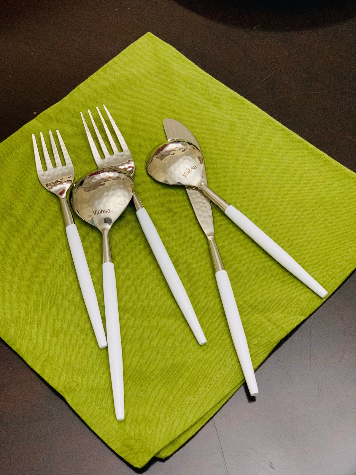 Set of stainless steel flatware with white handles including two forks, two spoons, and one butter knife displayed on a green cloth against a dark surface.