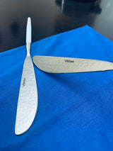 Two stainless steel butter knives with hammered texture and 'Vibhsa' engraving, one with a white handle, placed on a bright blue fabric surface.