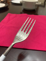 Close-up of a stainless steel fork with a hammered texture on the handle, resting on a bright pink cloth on a dark table with blurred white dishes in the background.