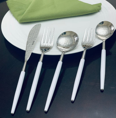 White and silver stainless steel flatware set with hammered mouth detailing laid out on dark surface