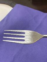 Close-up of a stainless steel fork with a hammered texture on the head, resting on a purple fabric surface with part of a white plate visible nearby.