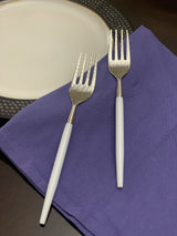 Two stainless steel dinner forks with hammered tines and smooth white handles resting on a purple cloth next to a white plate with a textured dark rim.
