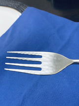 Close-up of a stainless steel dinner fork with a hammered texture on the mouth, resting on a blue fabric background with part of a white plate visible.