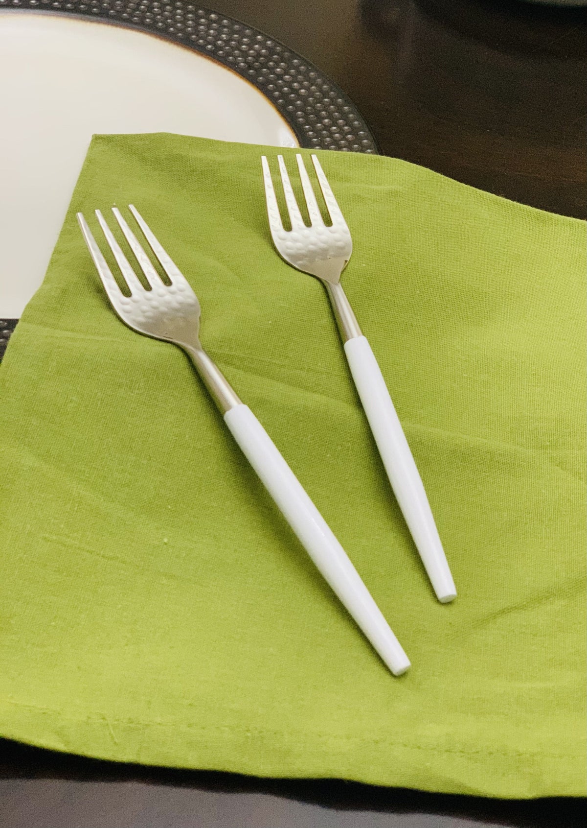 Set of six Vibhsa stainless steel dinner forks with hammered mouth and white handles arranged on a beige cloth