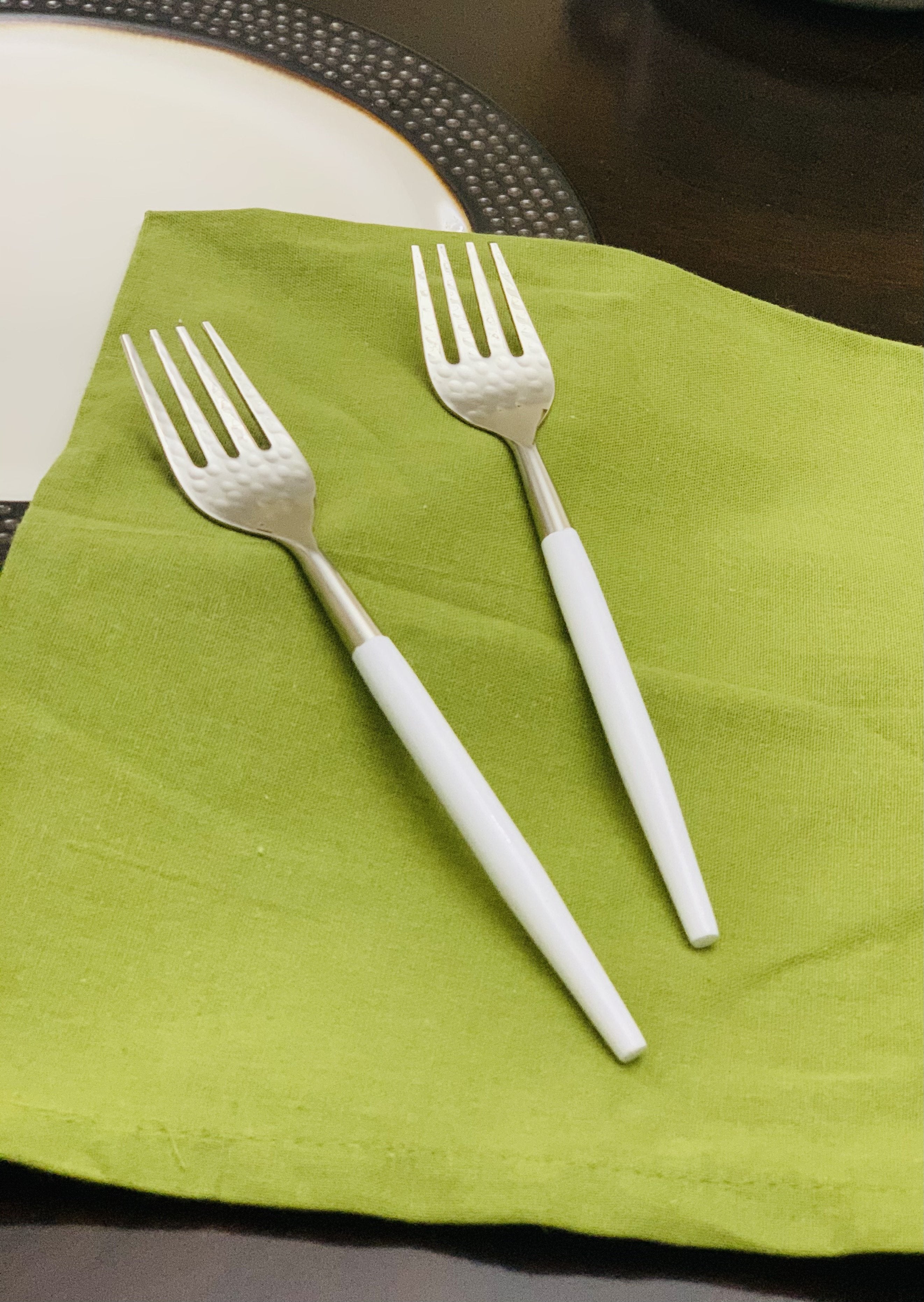 Set of six Vibhsa stainless steel dinner forks with hammered mouth and white handles arranged on a beige cloth