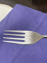 Close-up of a stainless steel fork with a hammered texture on the tines, resting on a blue fabric surface with a white plate partially visible.