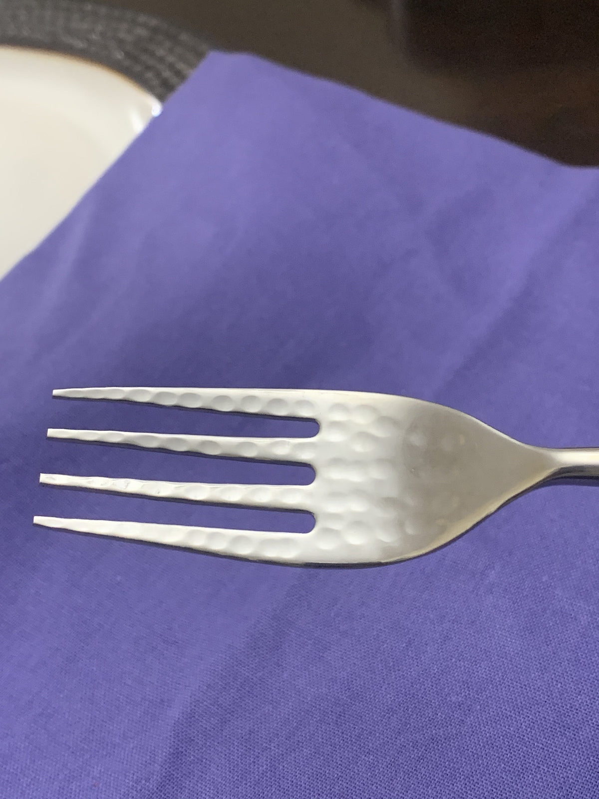 Close-up of a stainless steel fork with a hammered texture on the tines, resting on a blue fabric surface with a white plate partially visible.