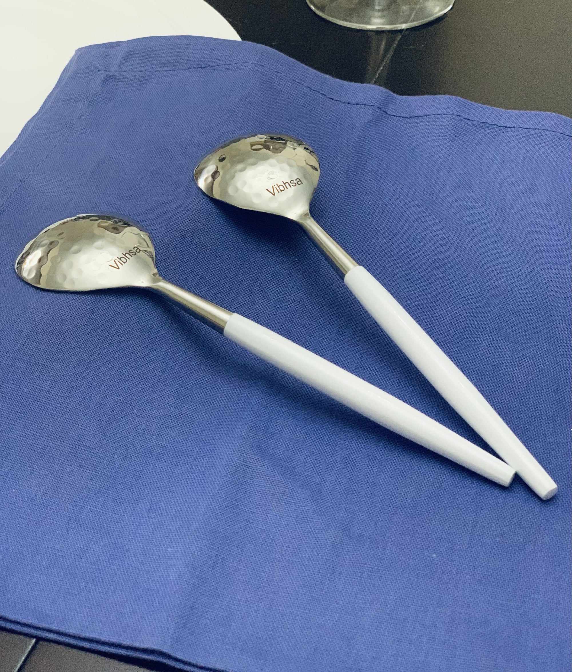 Vibhsa 6-piece stainless steel soup spoon set with hammered mouths and white handles arranged on a cloth background