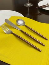 Three-piece hammered stainless steel flatware set with textured copper handles displayed on a bright yellow cloth against a dark wooden table.