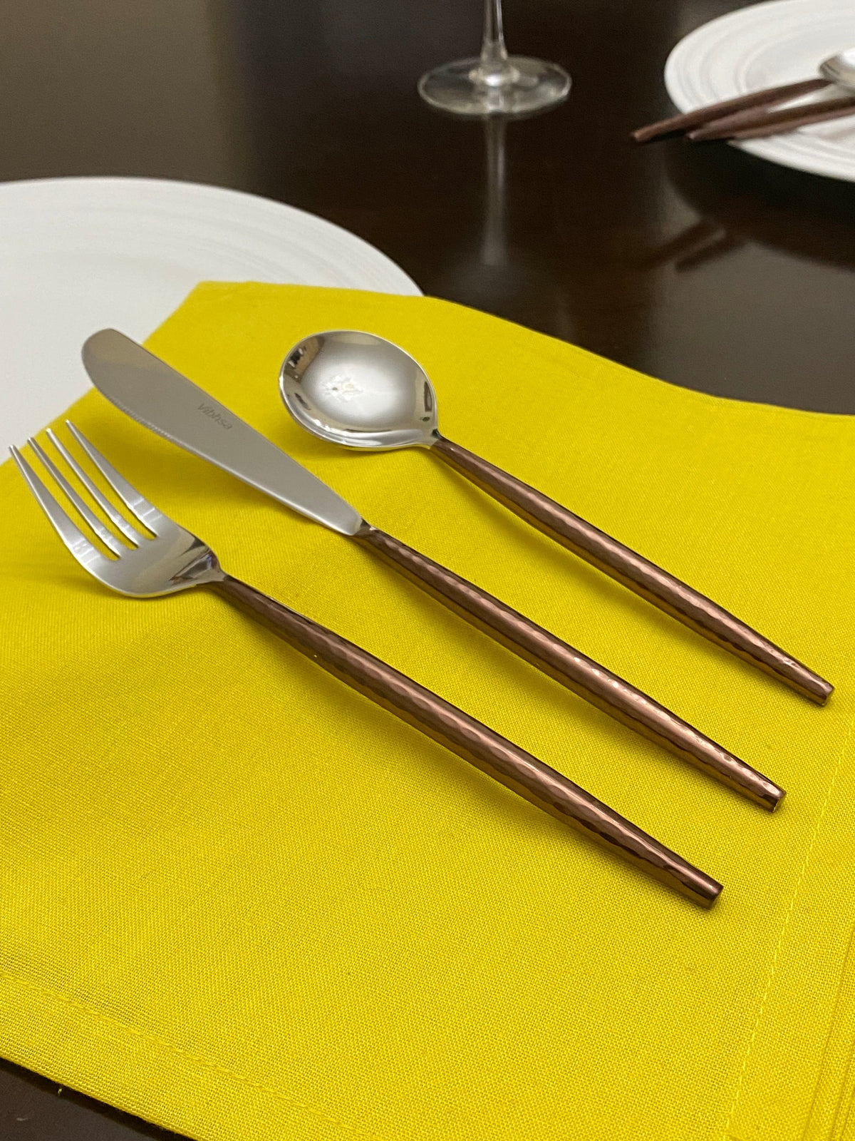Three-piece hammered stainless steel flatware set with textured copper handles displayed on a bright yellow cloth against a dark wooden table.