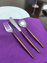 Fork, knife, and spoon with hammered copper-colored handles and shiny silver heads arranged on a purple cloth napkin beside a white plate on a dark table.
