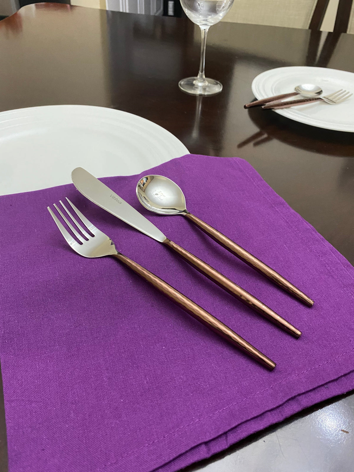 Fork, knife, and spoon with hammered stainless steel heads and dark wooden handles resting on a folded purple cloth on a dark wood table, with white plates and a wine glass in the background.