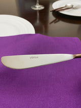Close-up of a butter knife with a serrated edge engraved with the name 'Vibhsa' resting on a purple cloth napkin, with white plates and glassware blurred in the background.