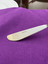 Stainless steel butter knife with serrated edge, engraved with 'Vibhsa', resting on a rich purple cloth beside a white plate on a dark wooden surface.