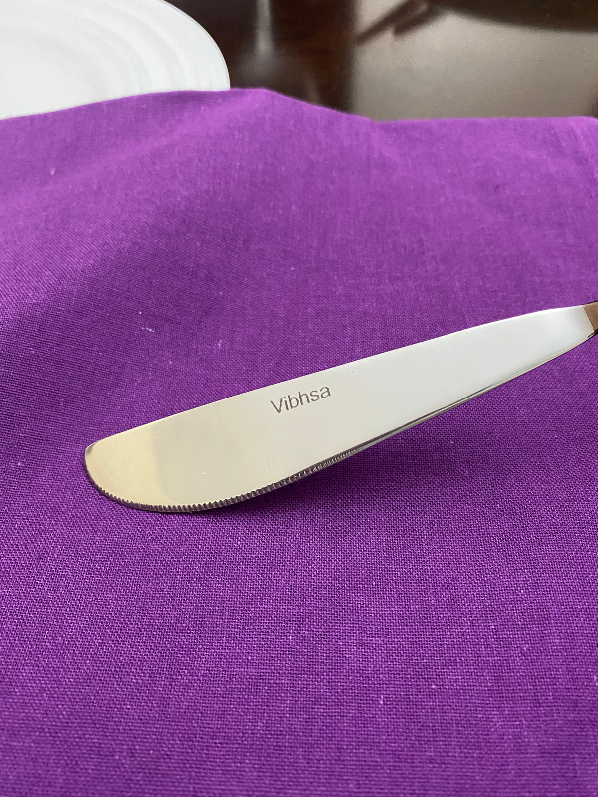 Stainless steel butter knife with serrated edge, engraved with 'Vibhsa', resting on a rich purple cloth beside a white plate on a dark wooden surface.