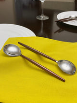 Two stainless steel spoons with long, slender brown handles resting on a bright yellow fabric napkin on a dark table, with white plates and a wine glass in the background.