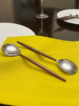 Two stainless steel round-bowled spoons with brown wooden handles resting on a bright yellow cloth, set on a dark table with white plates and a wine glass in the background.