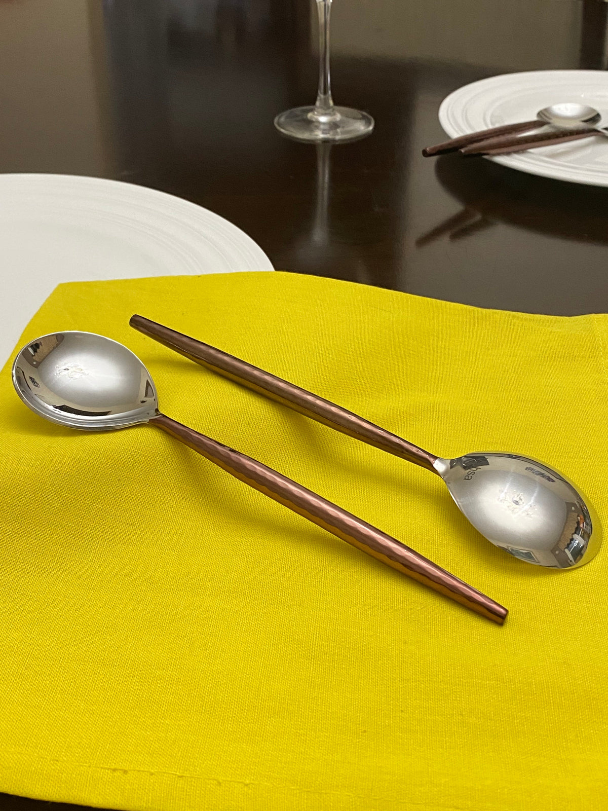 Two stainless steel round-bowled spoons with brown wooden handles resting on a bright yellow cloth, set on a dark table with white plates and a wine glass in the background.