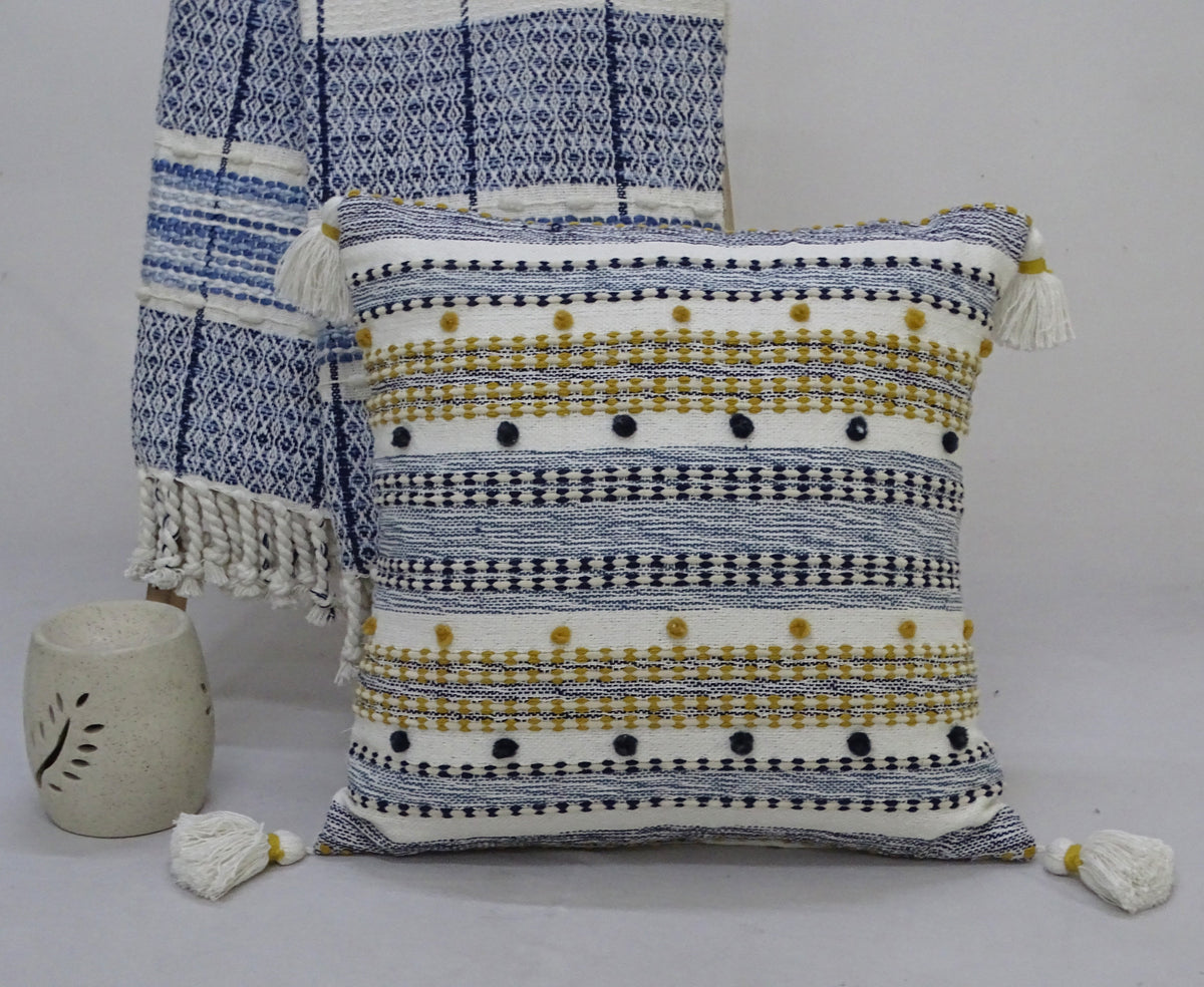 Square pillow with horizontal blue and mustard stripes, featuring small blue pom poms and white tassels on each corner, standing against a blue patterned textile.