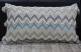 Rectangular chevron throw pillow in white and gray with blue accents, featuring braided zigzag patterns and tassels at each corner, positioned on a textured cushion.