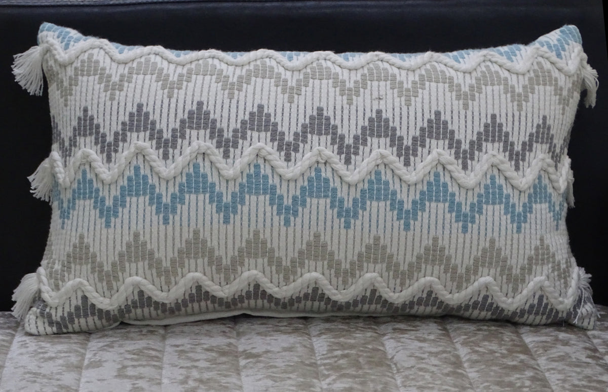 Rectangular chevron throw pillow in white and gray with blue accents, featuring braided zigzag patterns and tassels at each corner, positioned on a textured cushion.