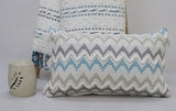 Rectangular chevron throw pillow in gray, blue, and beige with wavy edges, placed in front of a matching woven blanket on a gray surface with a speckled neutral-toned decorative holder nearby.