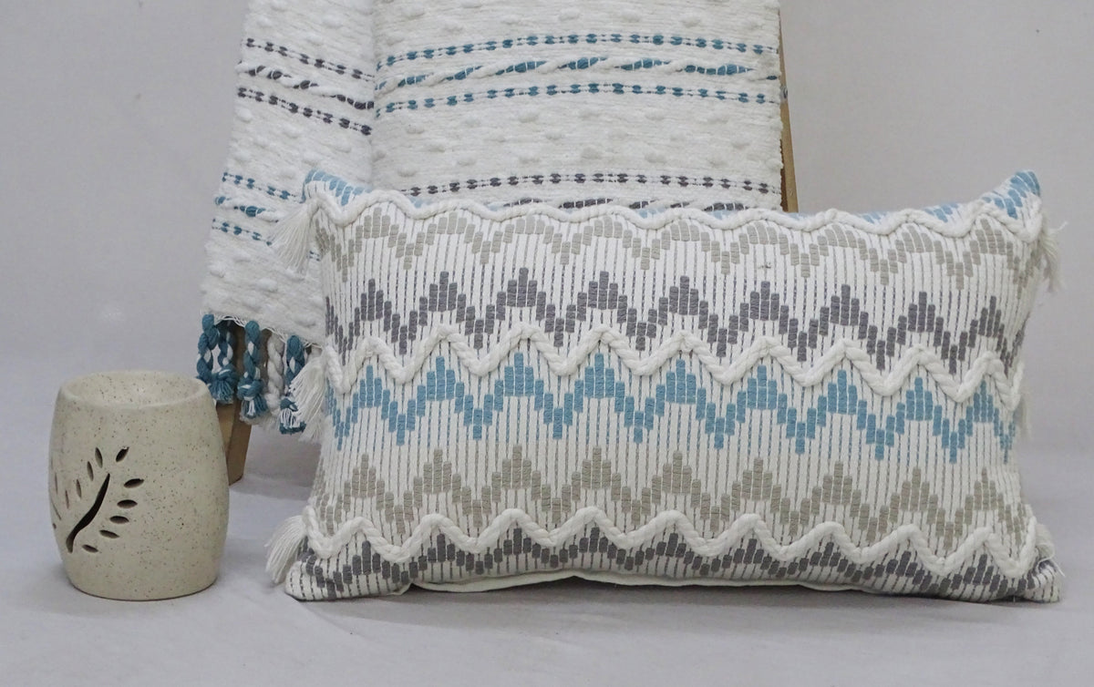Rectangular chevron throw pillow in gray, blue, and beige with wavy edges, placed in front of a matching woven blanket on a gray surface with a speckled neutral-toned decorative holder nearby.