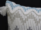 Close-up of a fabric cushion corner featuring a white braided zigzag with beige geometric patterns and blue rectangular accents above, with white tassels on the edge.