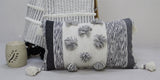 Rectangular throw pillow with white and gray striped pattern, decorated with large gray and white pom-poms and tassels on each corner, placed beside matching woven textile and candle holder.