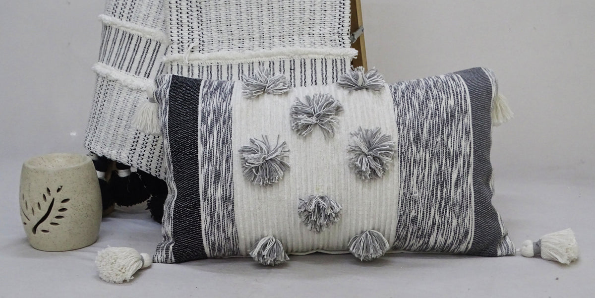 Rectangular throw pillow with white and gray striped pattern, decorated with large gray and white pom-poms and tassels on each corner, placed beside matching woven textile and candle holder.