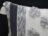 Close-up of a rectangular throw pillow with textured black and white stripes on one side and light gray pom-poms along one edge, featuring a white tassel at the corner.