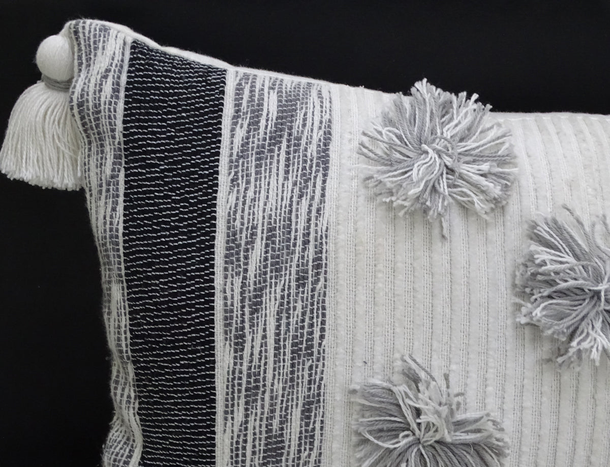 Close-up of a rectangular throw pillow with textured black and white stripes on one side and light gray pom-poms along one edge, featuring a white tassel at the corner.