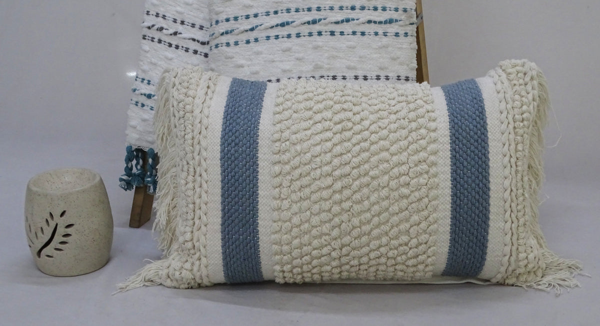 Rectangular decorative pillow with cream loop texture and two vertical blue stripes, featuring fringe on both ends, displayed in front of a folded textile with blue accents and a carved stone candle holder.