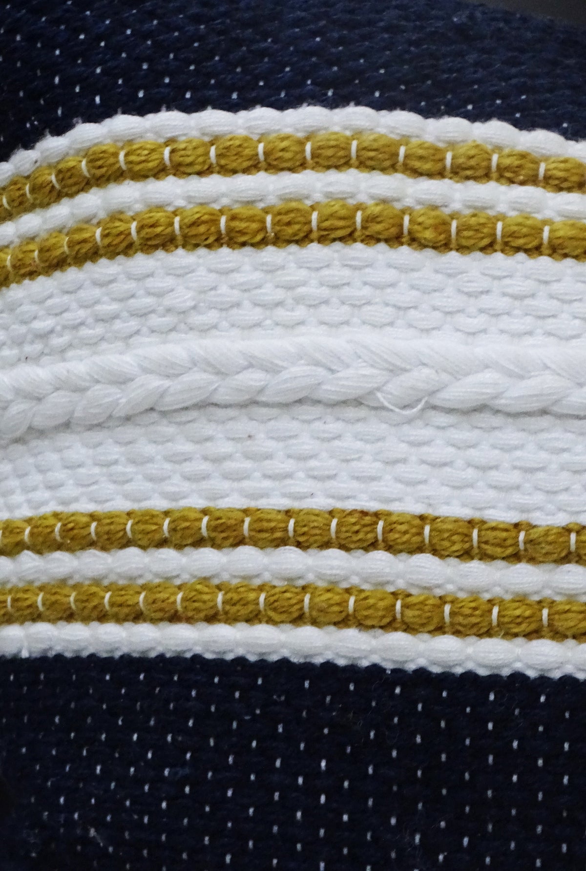 Close-up of a woven pillow with a textured white braid and mustard yellow stripes on a navy blue background.