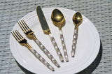 Twisted handle flatware set with gold-toned heads and textured silver handles displayed on a white plate against woven gray background, including forks, knife, and spoons.