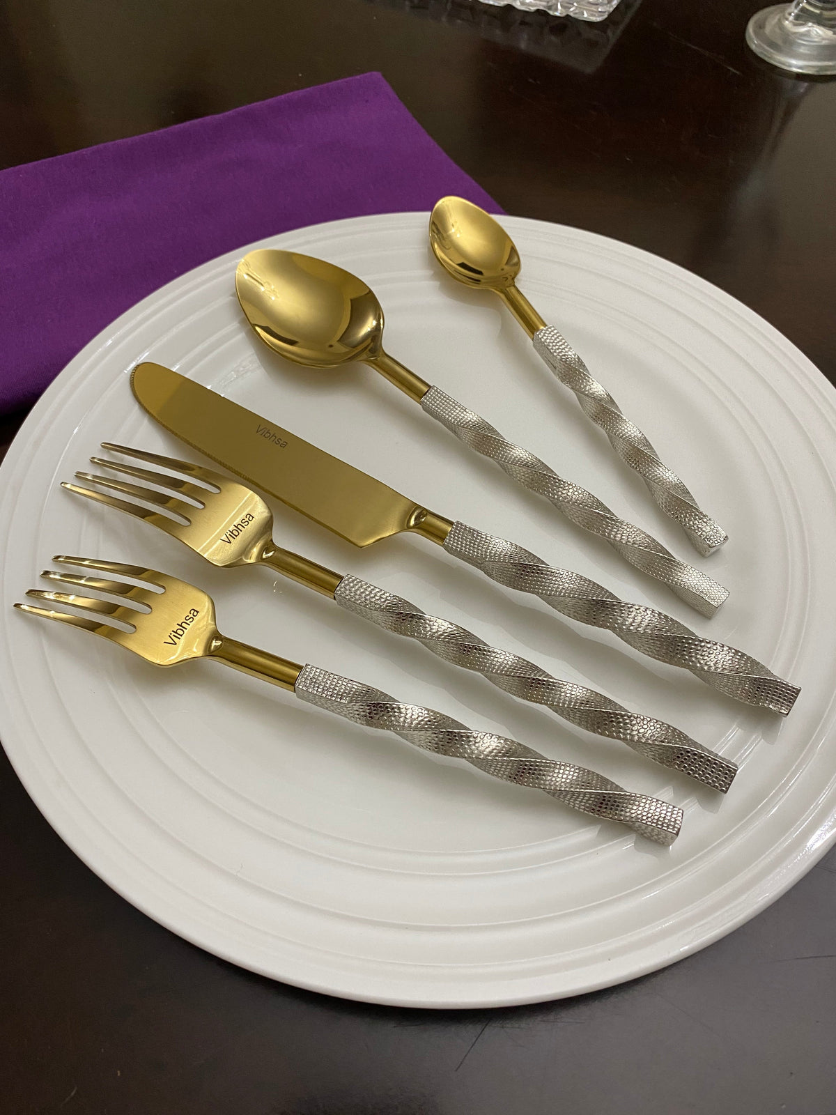Set of gold flatware with twisted silver handles displayed on a white plate, including forks, a knife, and spoons, with a purple cloth napkin nearby.