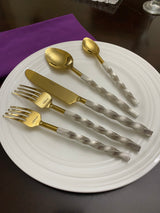 Gold flatware set with twisted textured silver handles arranged on a white plate, accompanied by a purple napkin on a dark wooden table.