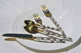 Golden flatware set with twisted textured silver handles, including two forks, one knife, and two spoons, arranged on a white round plate with concentric rings.