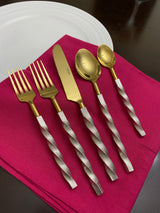 Five-piece golden stainless steel flatware set with twisted textured silver handles arranged on a magenta cloth over a white plate on dark wood.