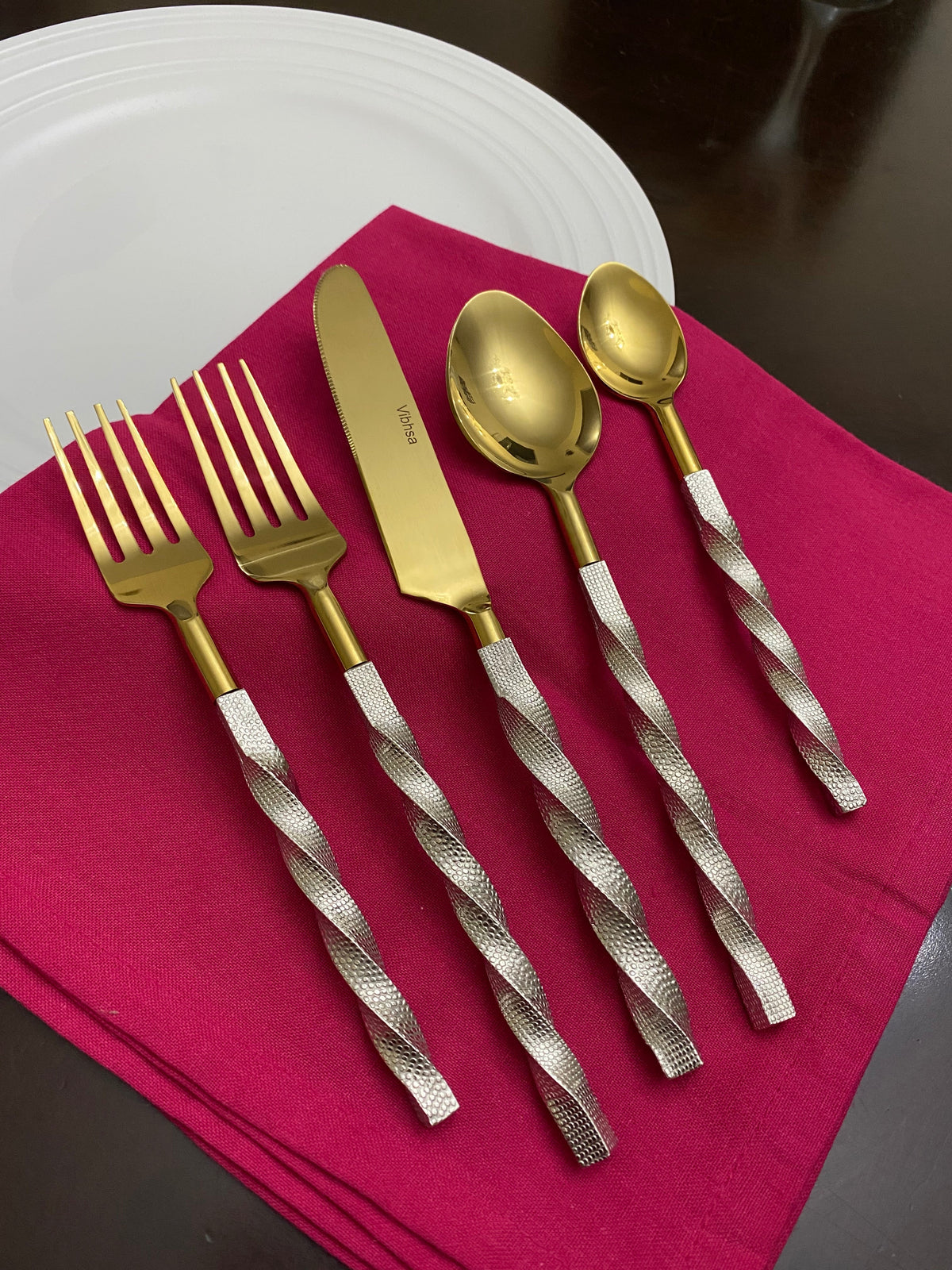 Five-piece flatware set with gold-toned heads and twisted silver textured handles, displayed on a magenta cloth atop a white plate and dark surface.
