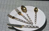 Golden stainless steel flatware set featuring twisted textured handles, including two forks, two spoons of different sizes, and a knife, arranged on a white plate over woven gray surface.