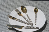 Golden stainless steel flatware set with twisted textured handles on a white round plate, placed on a gray woven surface.