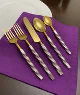 Set of five golden flatware pieces with twisted silver handles arranged on a purple cloth next to a white plate on a wooden surface.