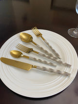 Set of golden stainless steel flatware with twisted textured silver handles arranged on a white round plate on a dark table.