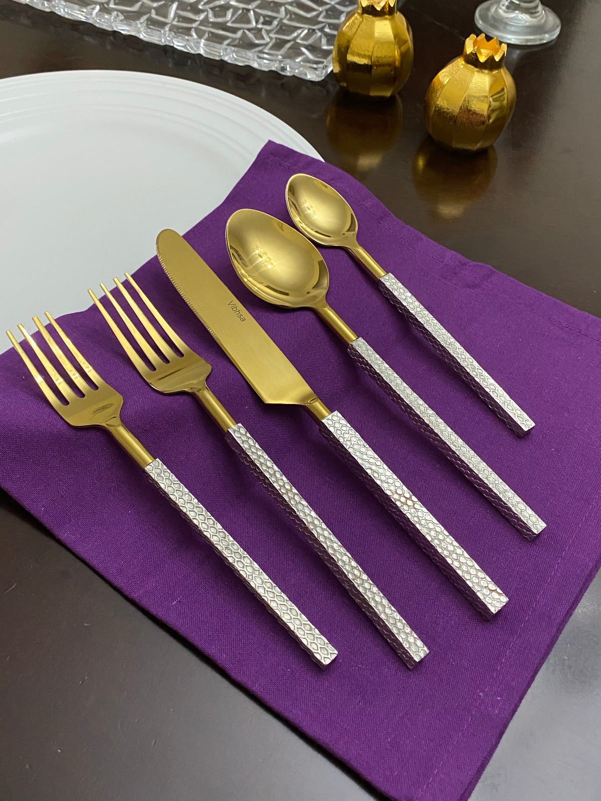 Golden stainless steel flatware set with textured silver handles, including two forks, a knife, and two spoons, placed on a purple cloth over a white plate.